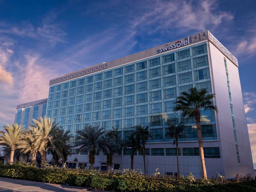 Un moderno edificio de hotel con un llamativo letrero de 'SWISSHOTEL', fachada de vidrio y rodeado de palmeras.