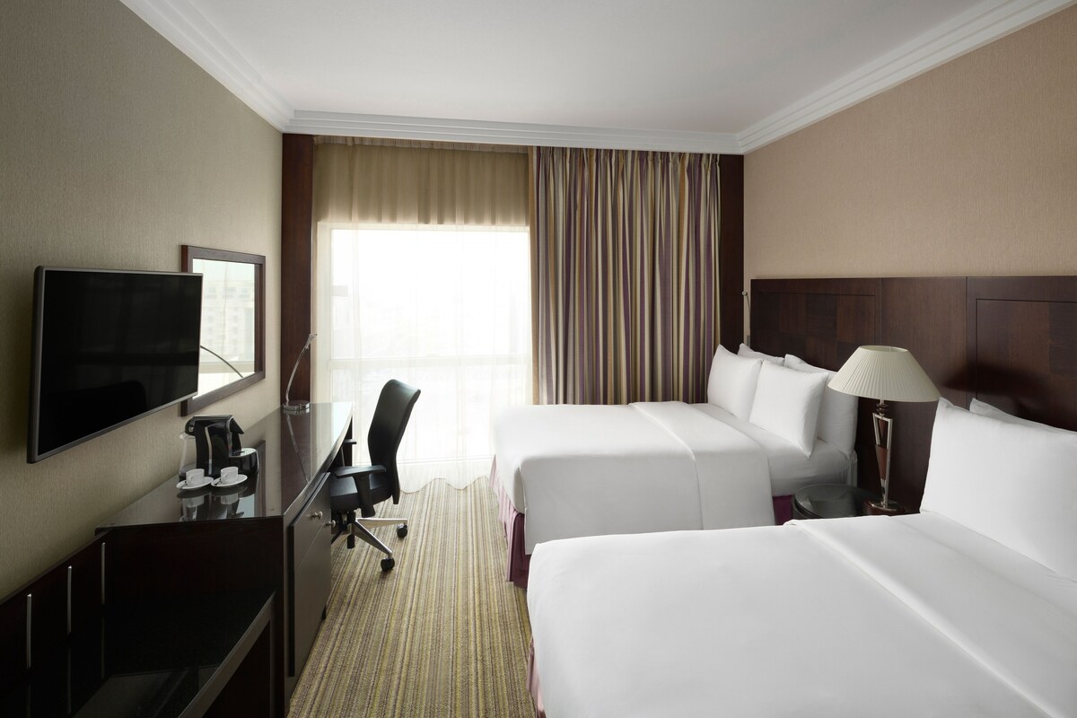 Une chambre d’hôtel avec deux lits, un bureau avec une télévision, une chaise et une fenêtre avec des rideaux.