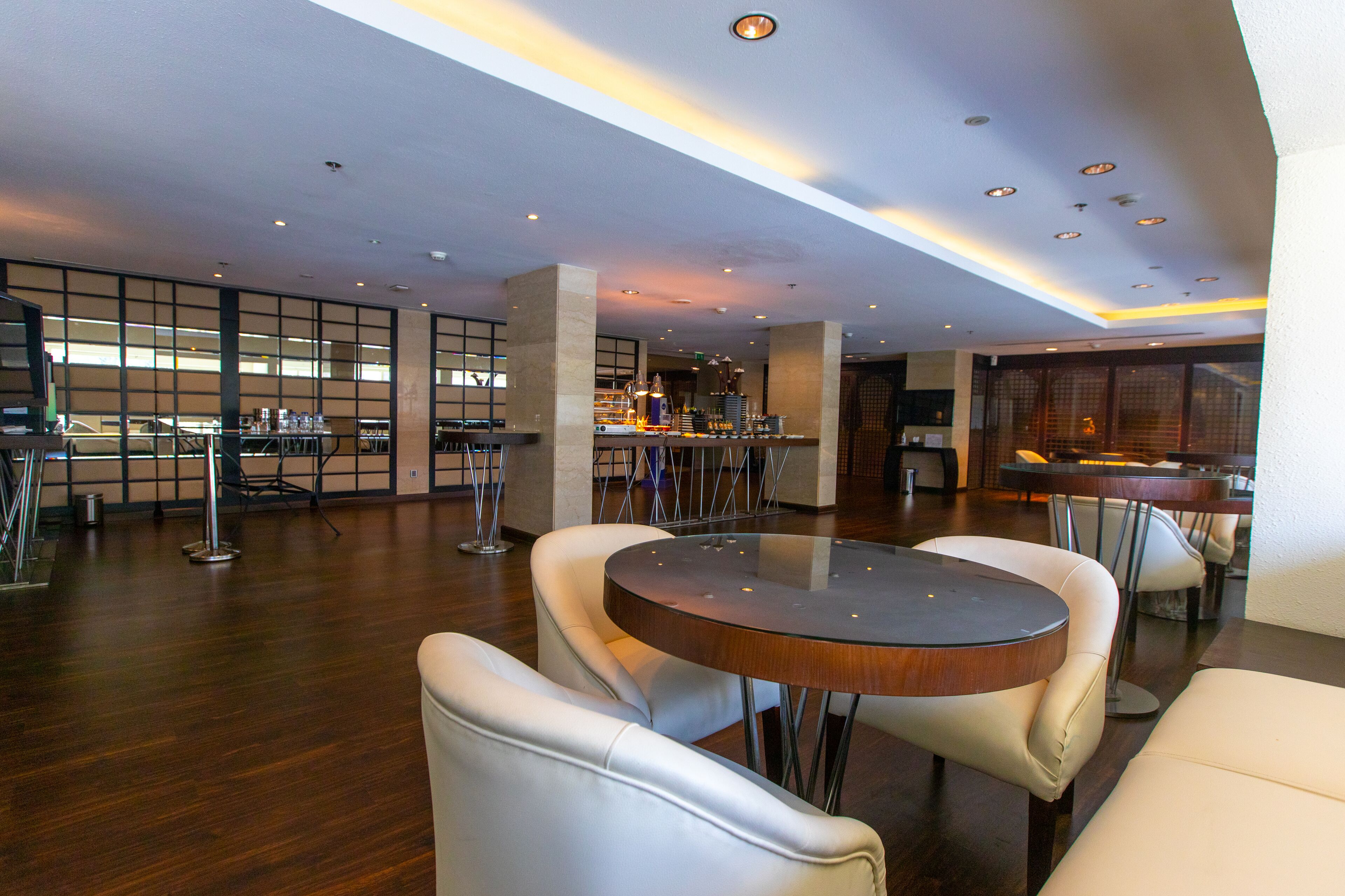 Um hall de hotel moderno com uma mesa de madeira redonda cercada por cadeiras de couro branco, área de bar ao fundo e um layout espaçoso com grandes janelas.