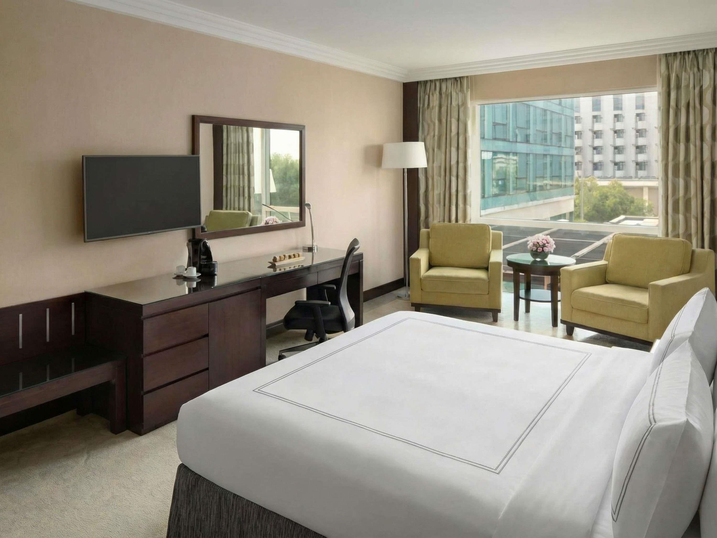 A hotel room with a bed, a desk, two chairs, a TV, and a window with a view of buildings.