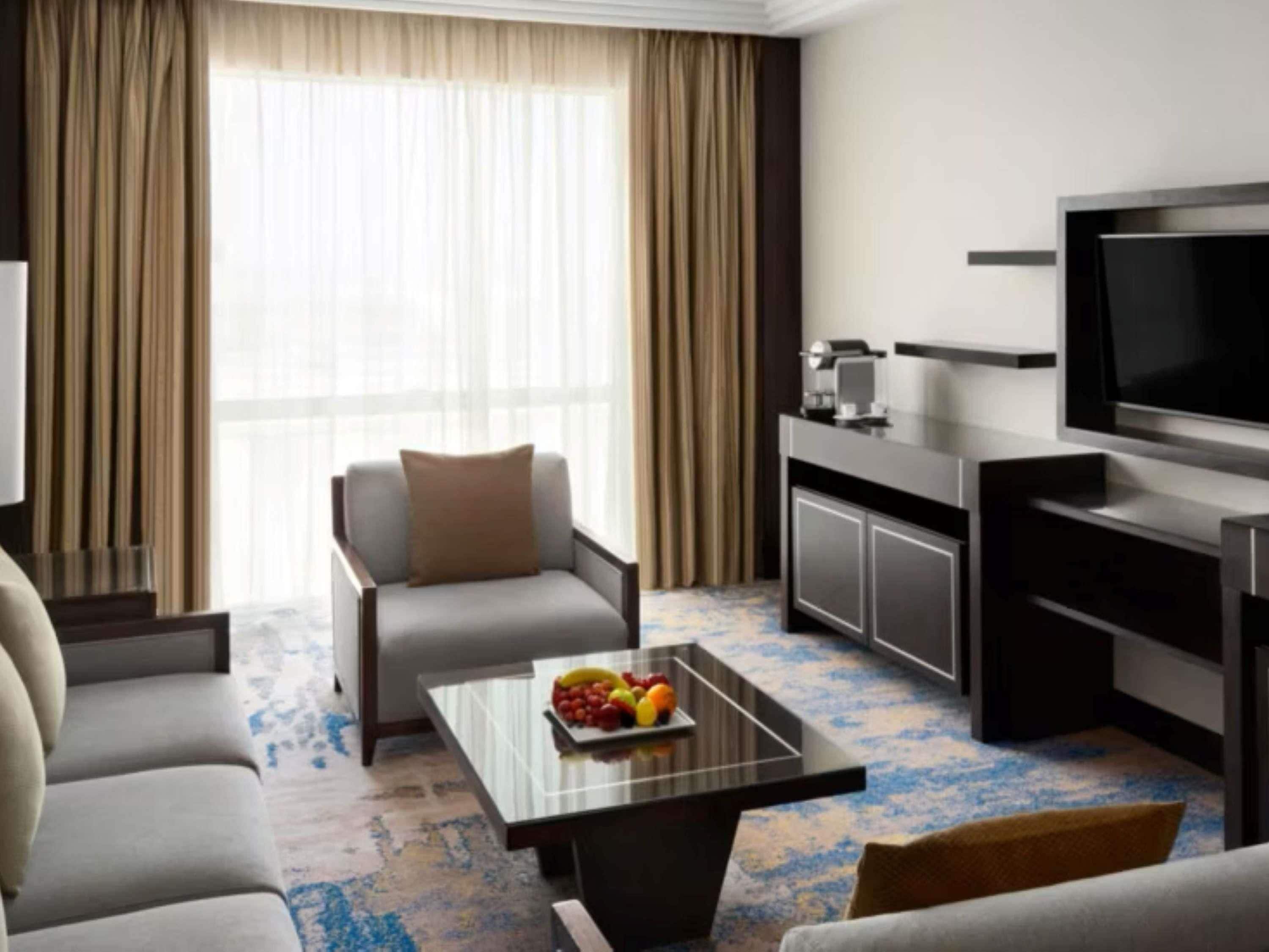 Une chambre d’hôtel moderne équipée d’un canapé, d’une table basse avec des fruits, d’une télévision et d’un mini-bar.