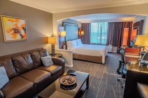 Una habitación de hotel con un sofá de cuero marrón, una cama con ropa blanca, un escritorio con lámpara y un ventanal con cortinas.