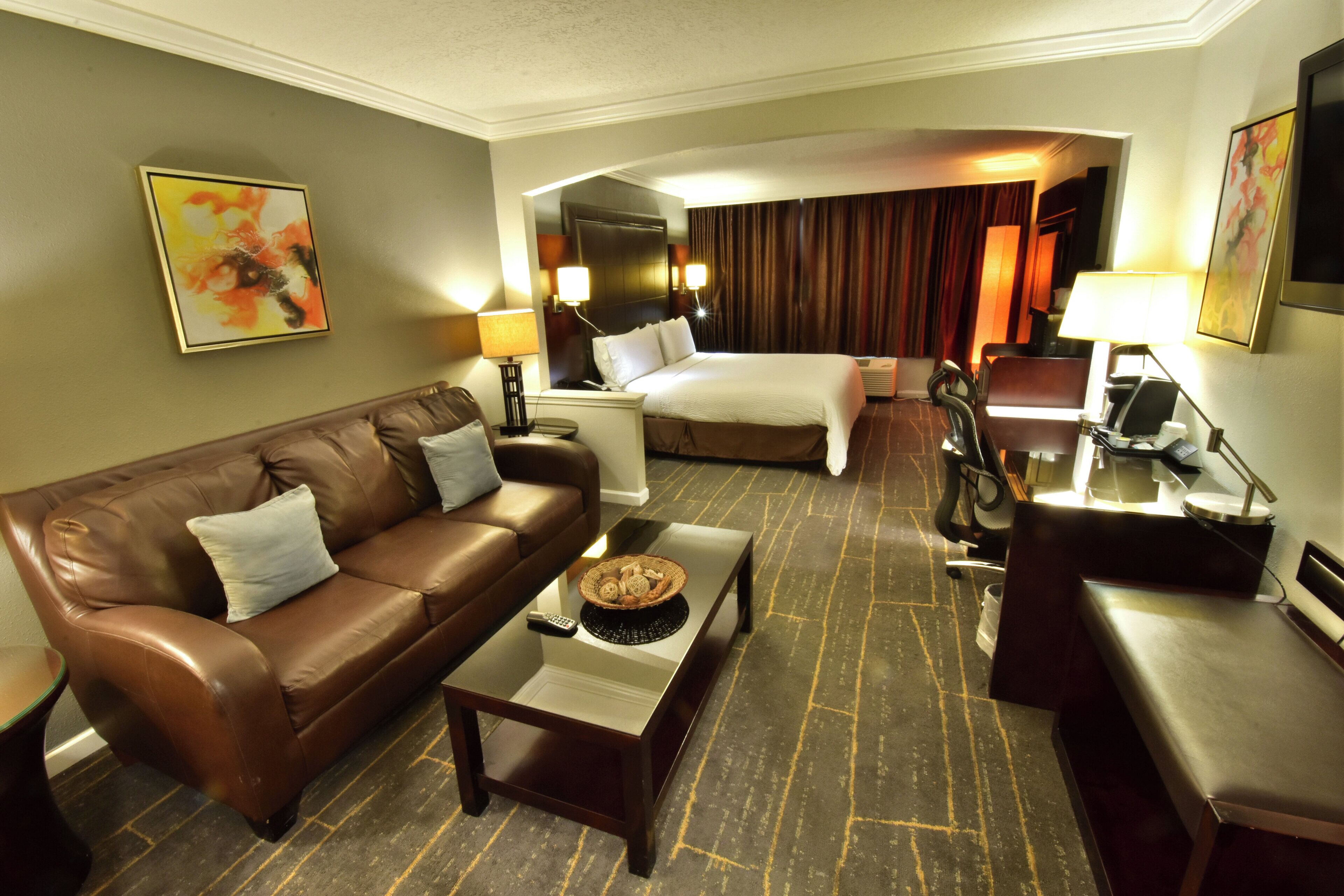 A hotel room with a brown leather sofa, a bed with white bedding, a desk with a lamp, and a painting on the wall.