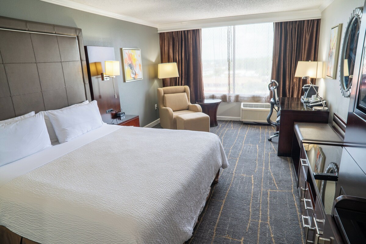 A hotel room with a large bed, a desk with a chair, a small table, a lamp, a mirror, and a window with curtains.