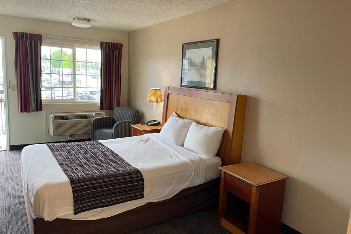 Una habitación de hotel con cama, mesita de noche, lámpara, silla y ventana con cortinas.
