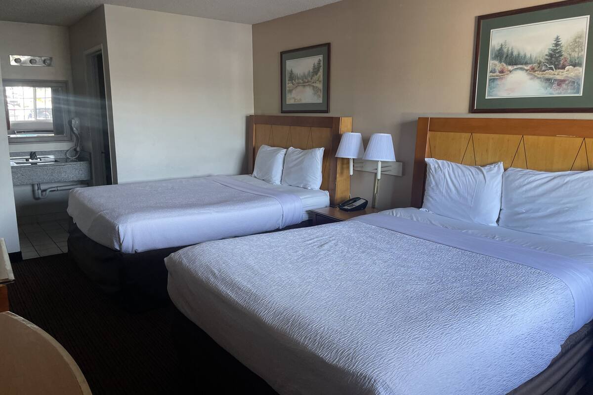 A hotel room with two beds, a wooden headboard, a nightstand with a lamp, a painting on the wall, and a bathroom visible through an open door.