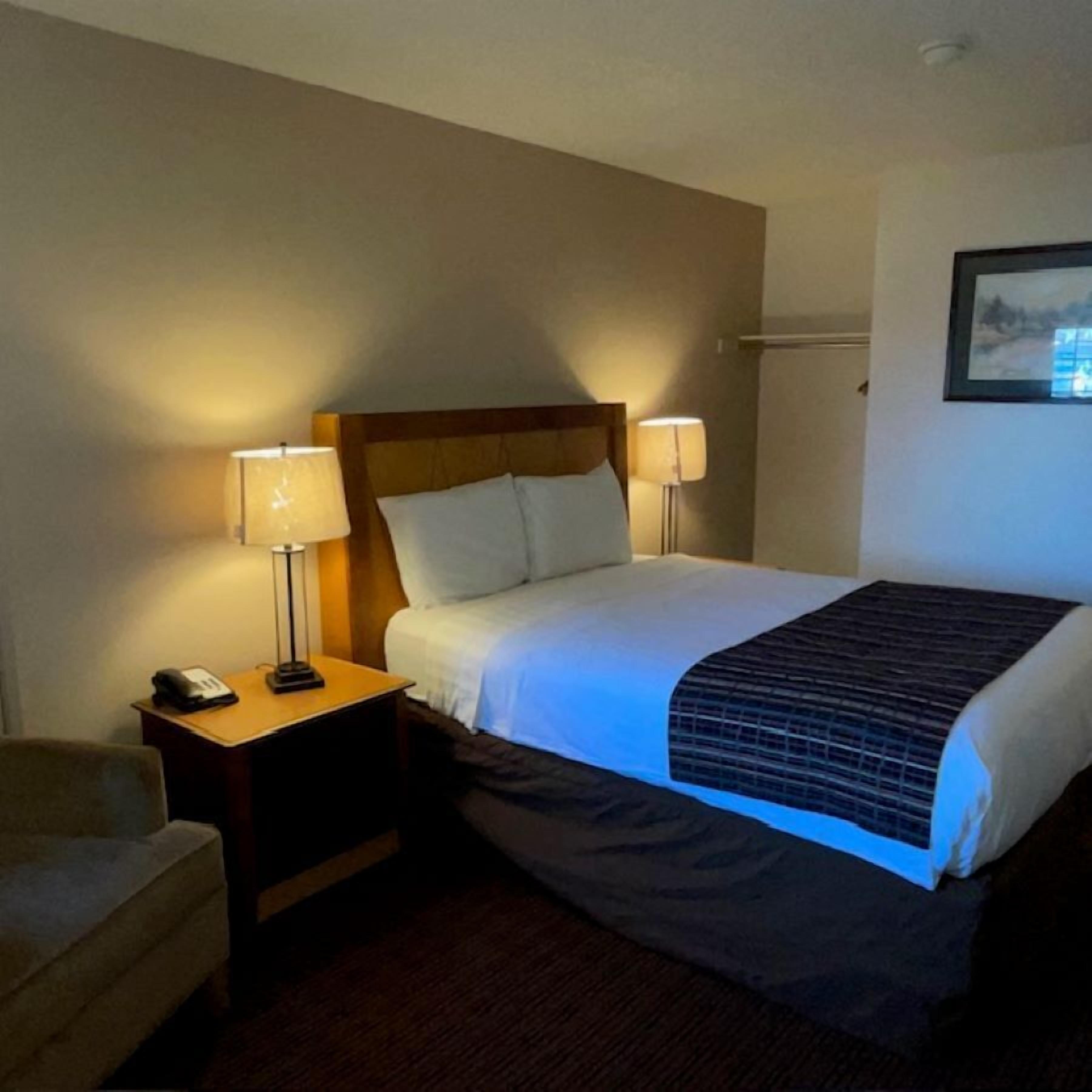 A hotel room with a bed, two bedside lamps, a nightstand with a phone, and a chair.