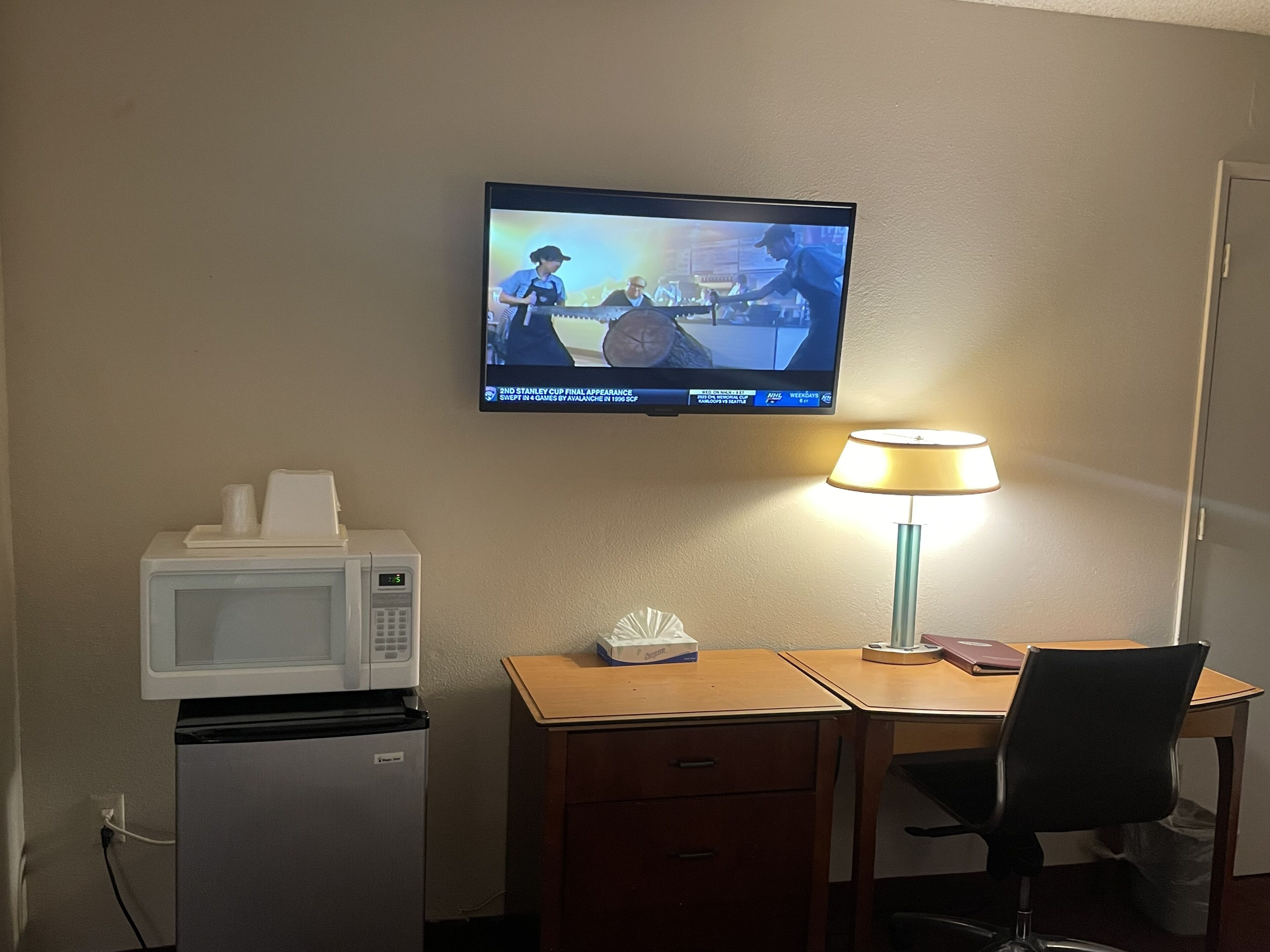 Hotelkamer met een aan de muur bevestigde televisie waarop een sportevenement wordt getoond, een magnetron, een kleine koelkast, een bureau met een lamp en een stoel.