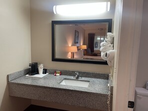 A hotel bathroom with a granite countertop, a sink, a mirror, a towel dispenser, and a toilet.