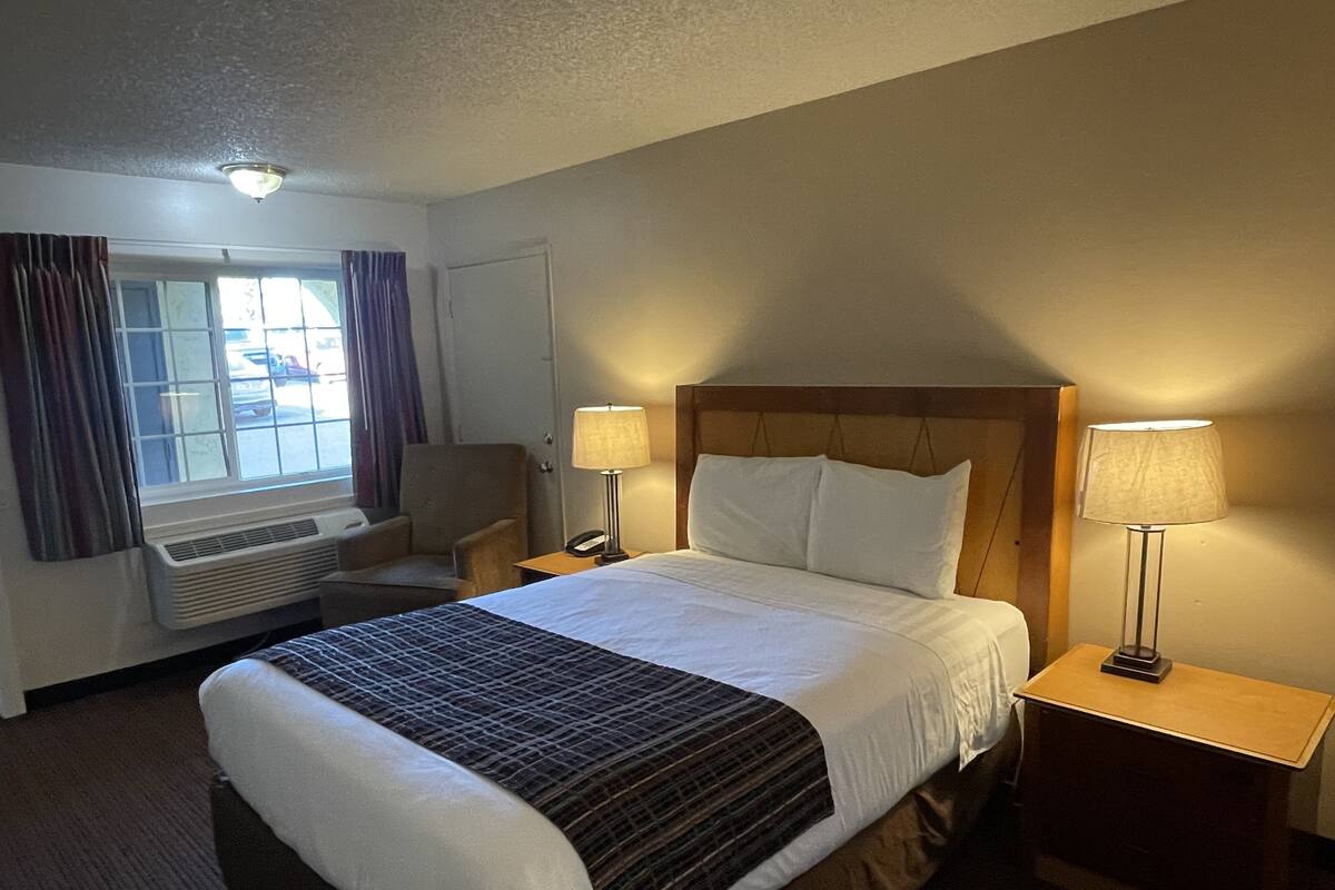 A hotel room with a large bed, two bedside lamps, a chair, a window with curtains, and a thermostat.