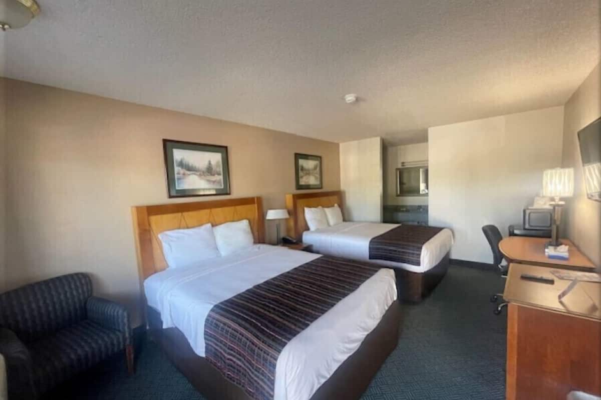 A hotel room with two beds, a desk, a chair, and a small kitchenette.