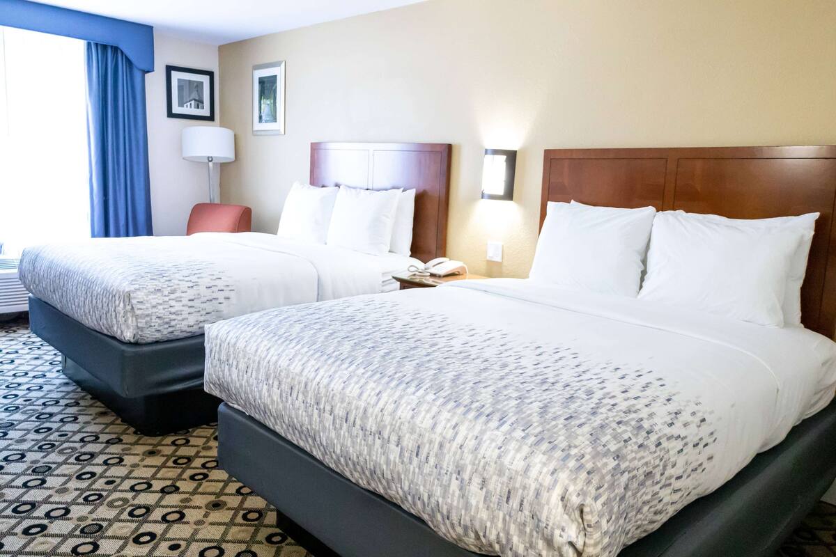Habitación de hotel con dos camas, un escritorio, una silla, una lámpara y una ventana con cortinas.