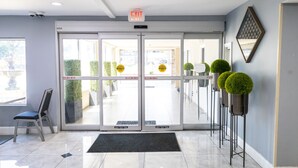 Entrada de hotel con puerta de vidrio, una silla, felpudo y plantas en macetas.