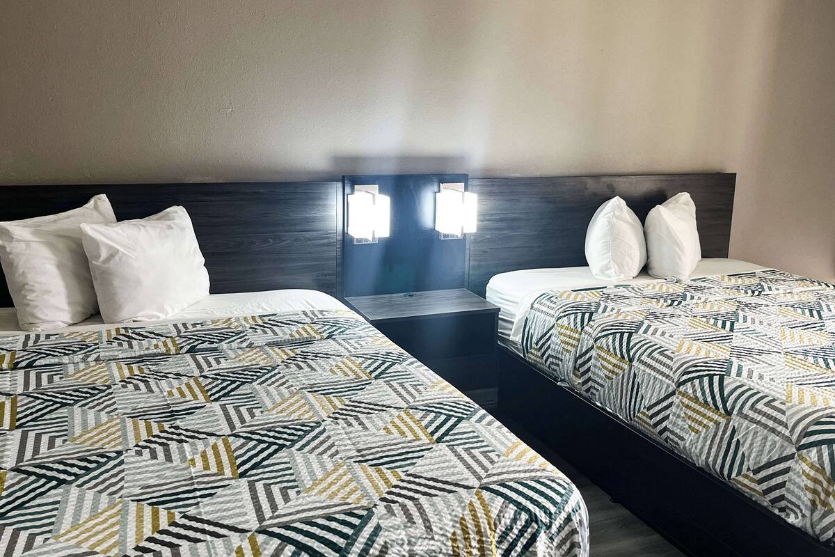 A hotel room with two beds, each with a patterned bedspread, a dark headboard, and a nightstand with a lamp.
