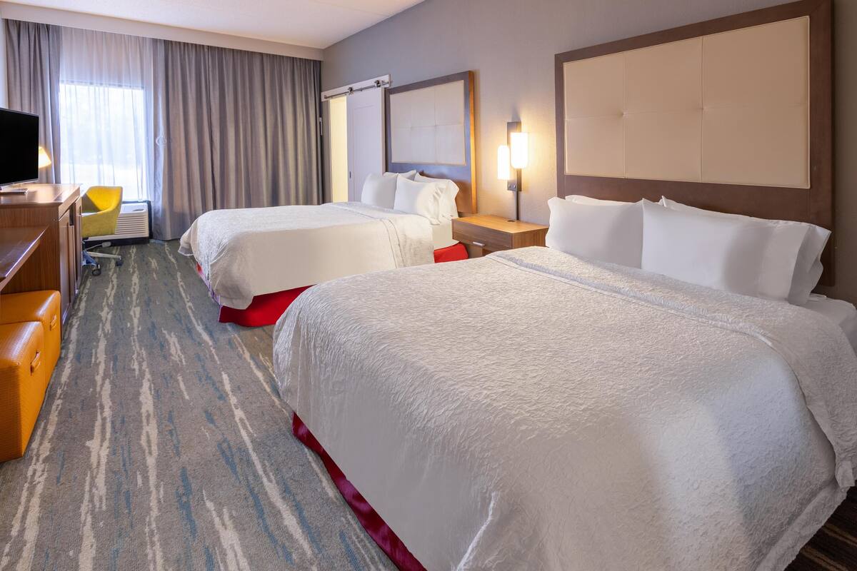 Habitación de hotel con dos camas, un escritorio con silla, un televisor y un ventanal con cortinas.