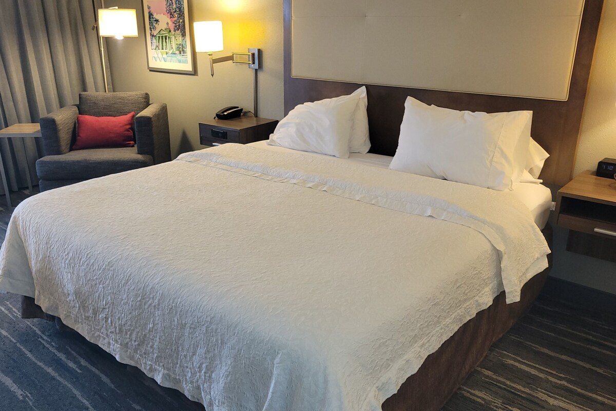 A hotel room with a large bed, a chair, a nightstand, a lamp, and a painting on the wall.