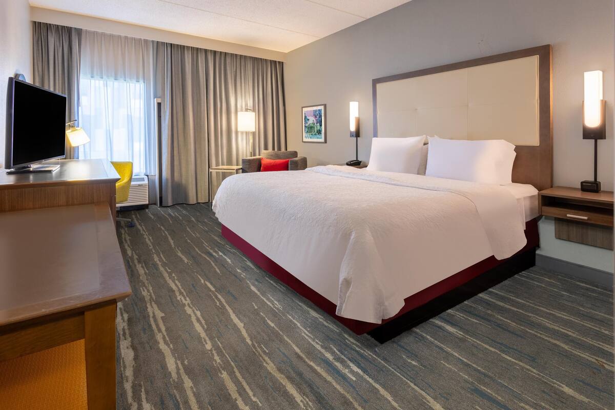 Habitación de hotel con una cama grande, un escritorio con televisor, una silla y dos lámparas de noche.