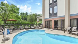 A swimming pool with lounge chairs and umbrellas, surrounded by trees and a building with large windows. - Hampton Inn Commerce (Commerce)