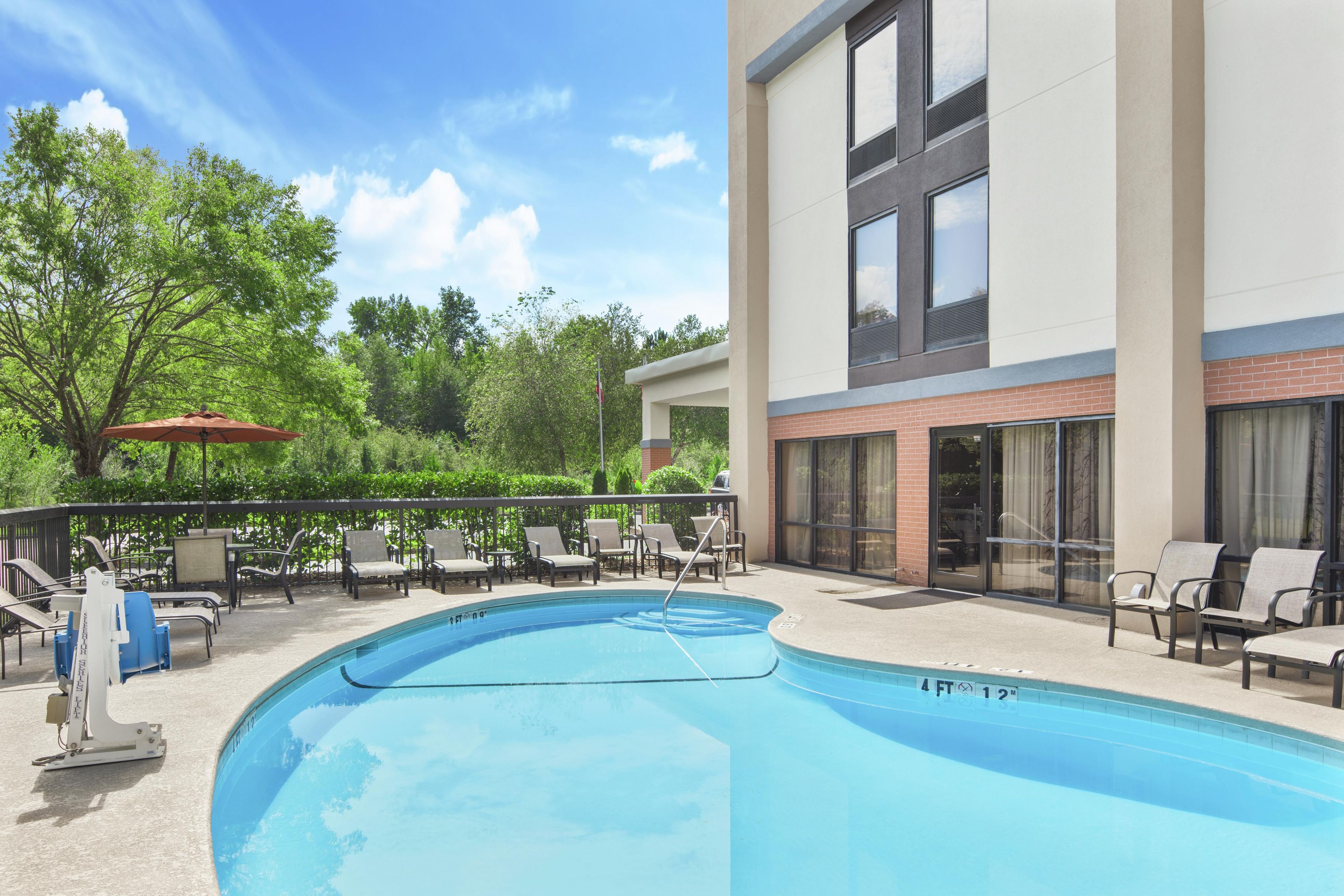 A swimming pool with lounge chairs and a red umbrella, surrounded by trees and a building with large windows.