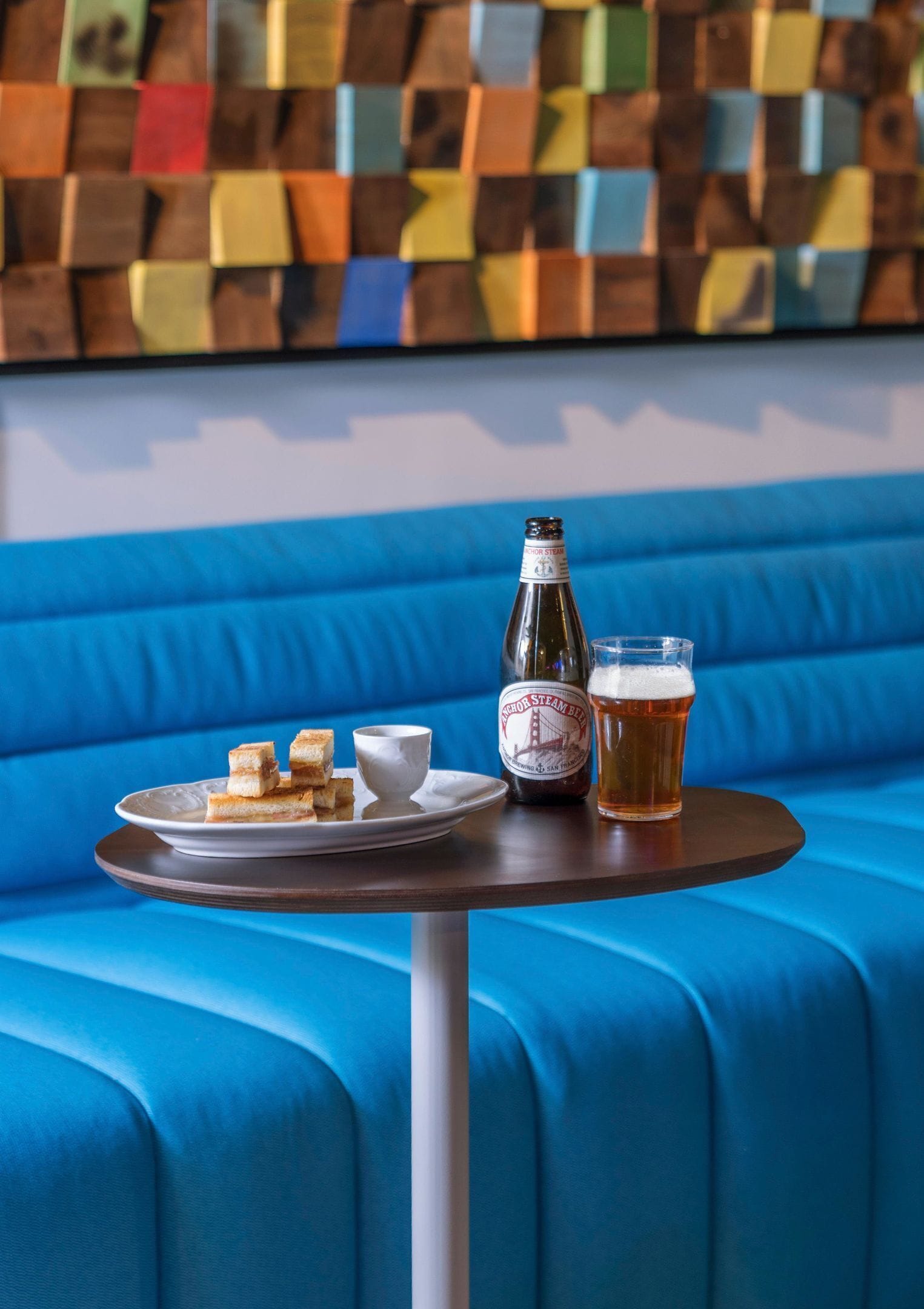 A round table with a bottle and a glass of beer, a plate with cubes and a cup, and a blue upholstered bench.