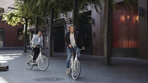 Deux personnes à vélo circulent dans une rue de la ville, avec des bâtiments en arrière-plan.