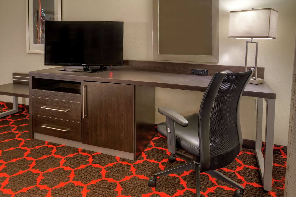 A hotel room with a desk, a television, a chair, and a lamp.