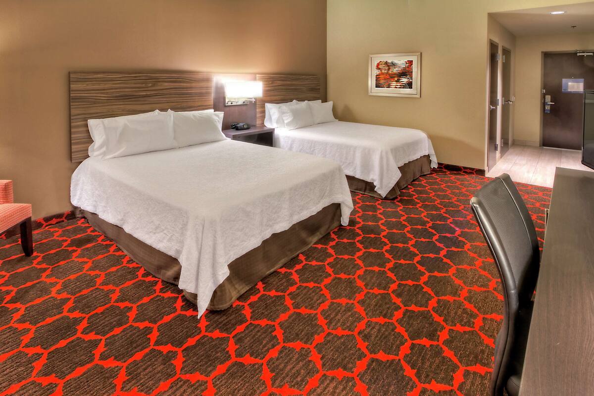 A hotel room with two beds, a desk, a chair, and a patterned carpet.