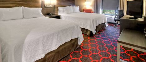 A hotel room with two beds, a TV, a desk, and a window with curtains.
