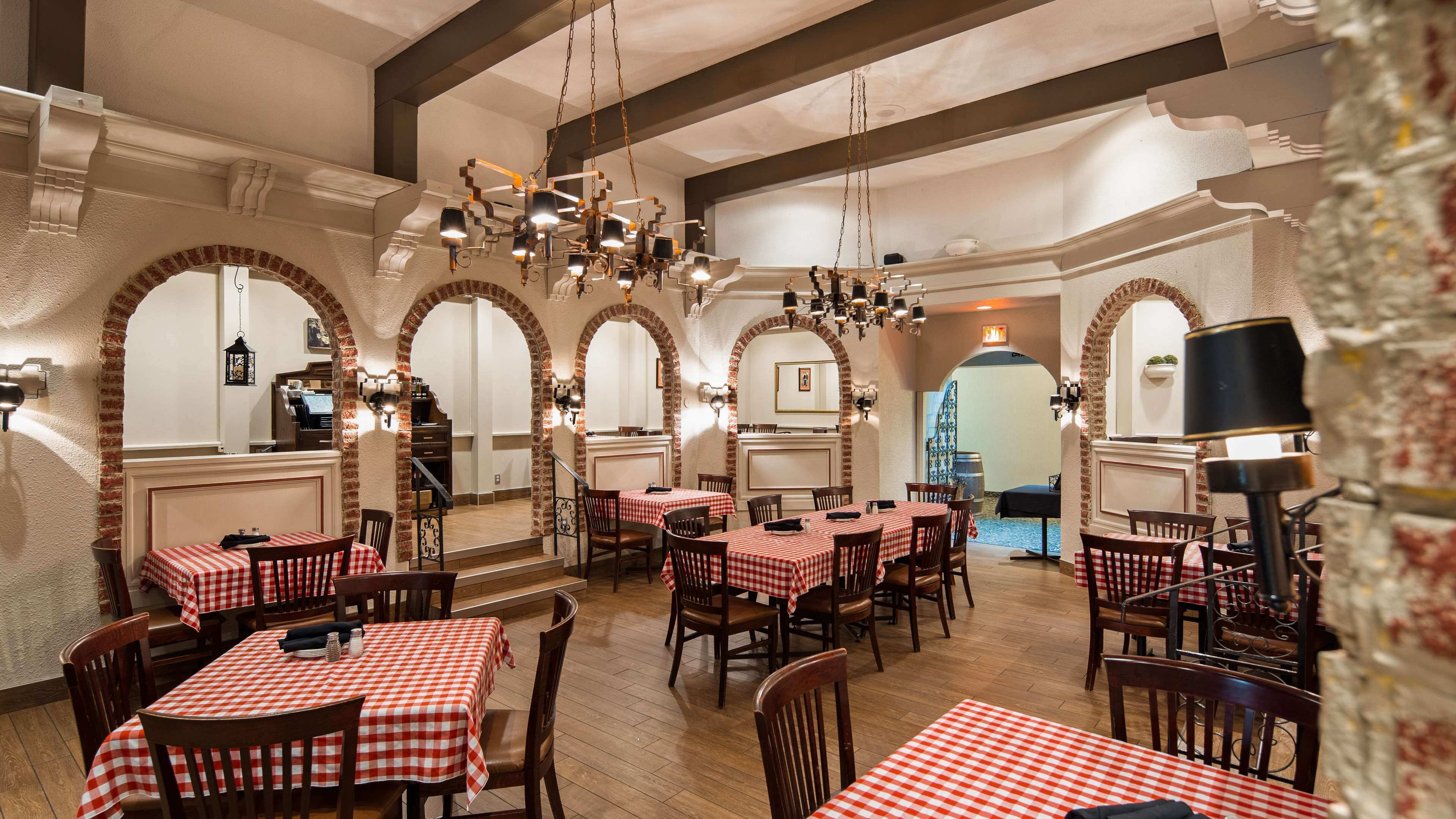 A restaurant with checkered tablecloths, wooden chairs, and brick arches. Tables are arranged in a spacious layout with hanging lights above.