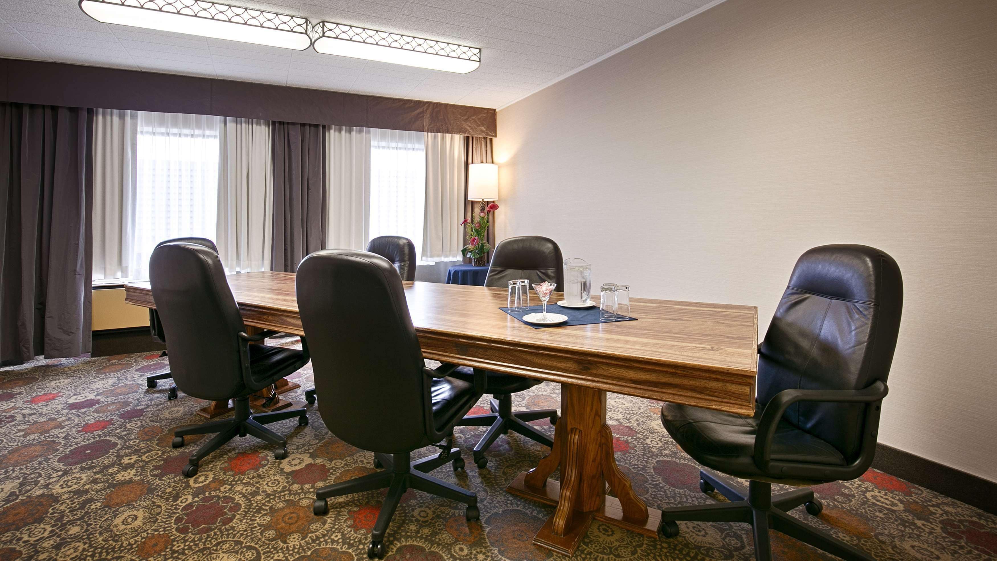 Un salón de conferencias con una mesa de madera, cuatro sillas de oficina negras, una alfombra estampada, un ventanal con cortinas y una lámpara de mesa.