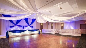 Un salón de banquetes con cortinas blancas, un escenario con fondo azul y filas de sillas blancas.