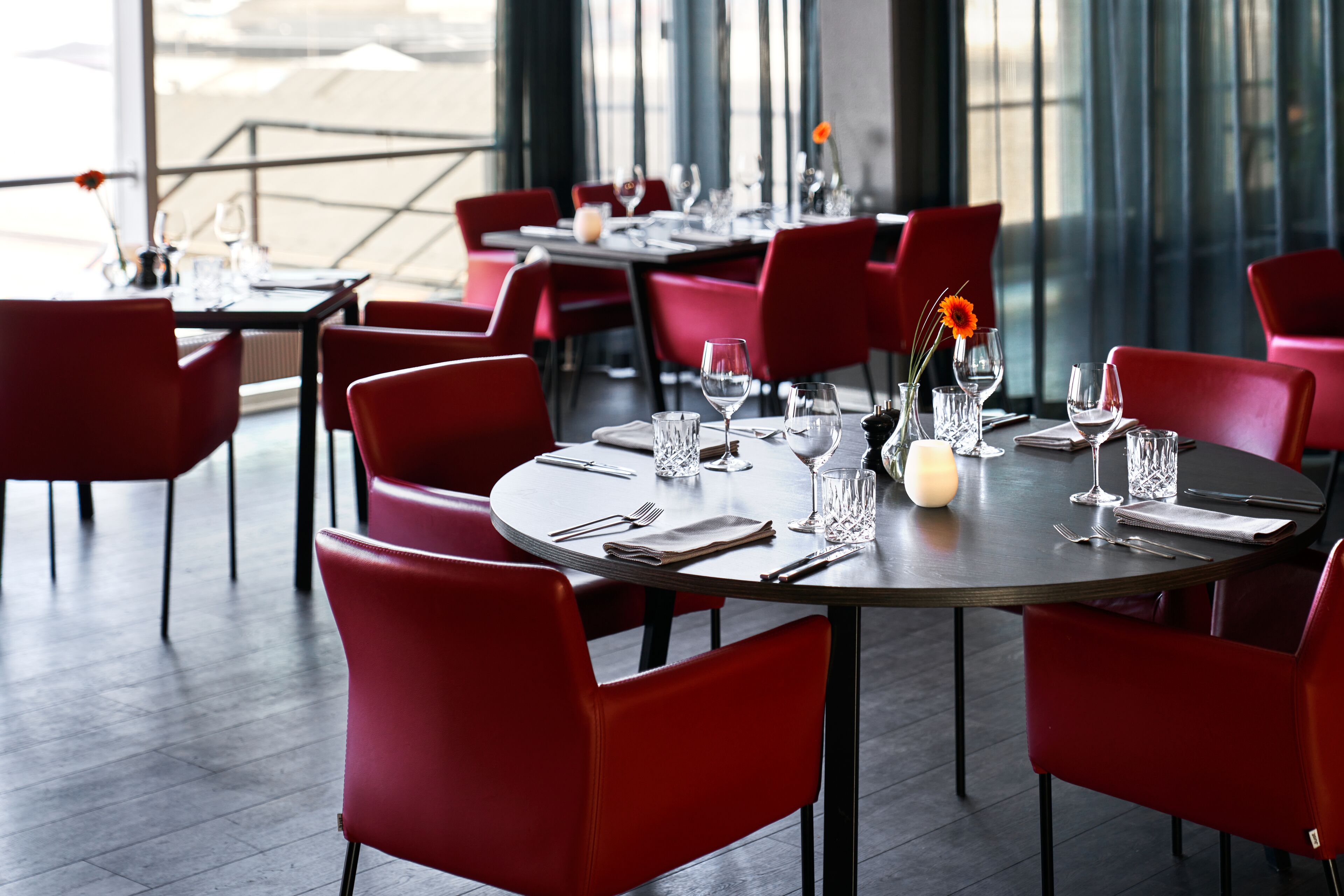 A restaurant with round tables and red chairs, set with wine glasses, cutlery, and a vase with a flower.