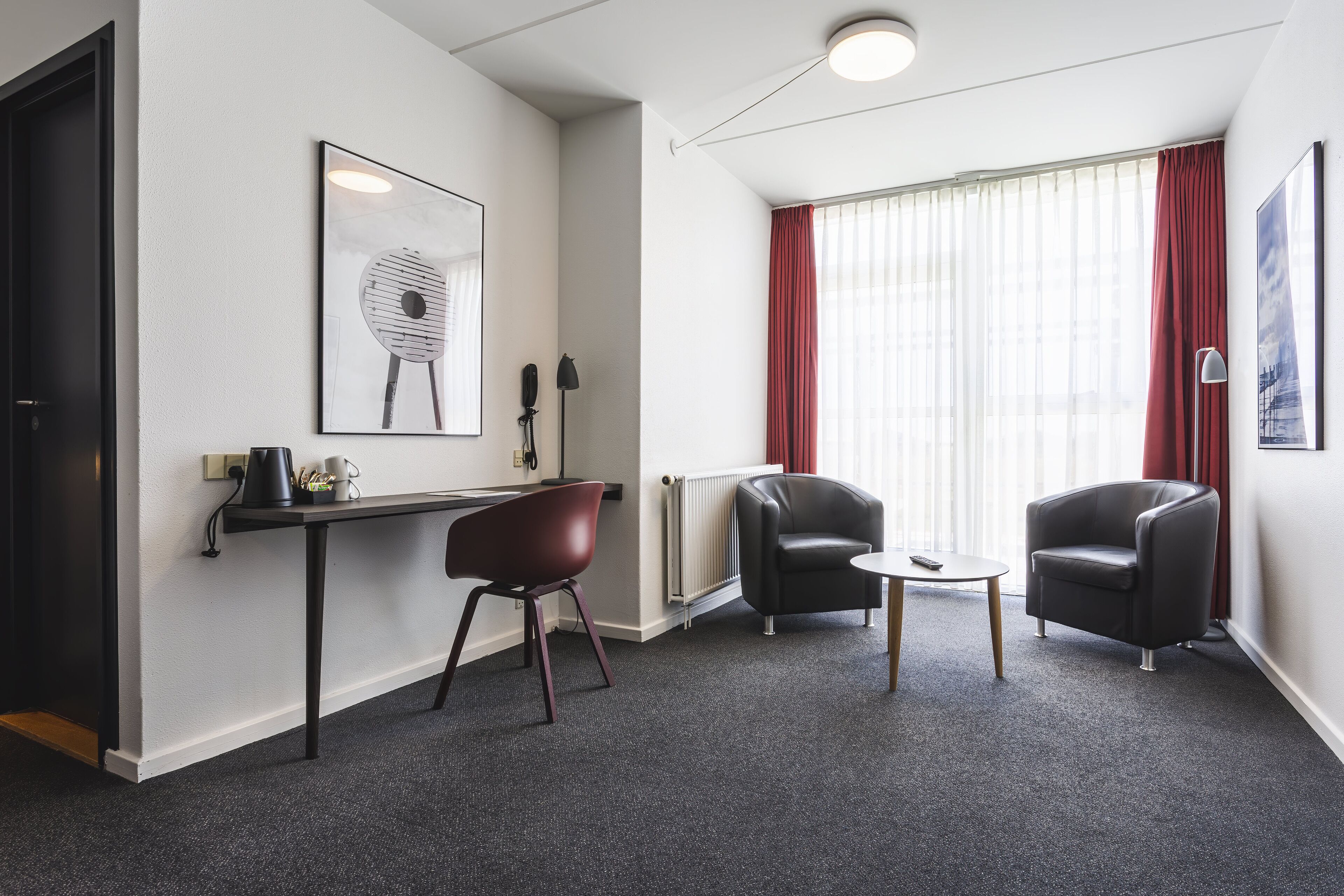 A modern hotel room with a desk, two chairs, a radiator, and a large window with red curtains.