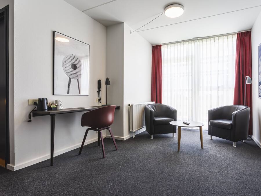 Una habitación de hotel moderna con un escritorio, dos sillas, un radiador y un ventanal grande con cortinas rojas.