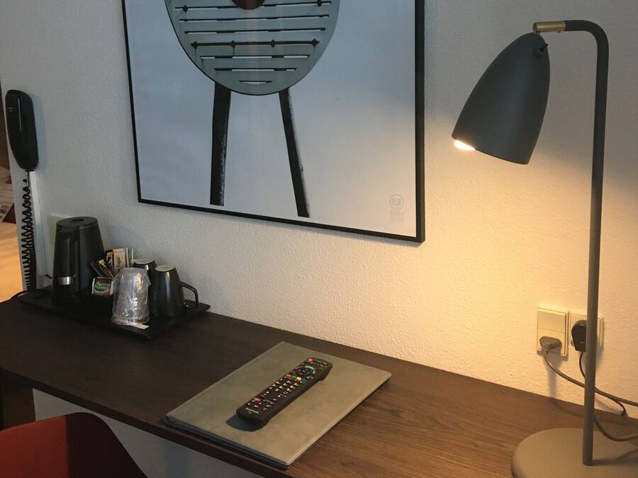 A hotel room with a desk, a red chair, a framed artwork, a desk lamp, a coffee maker, and a remote control.