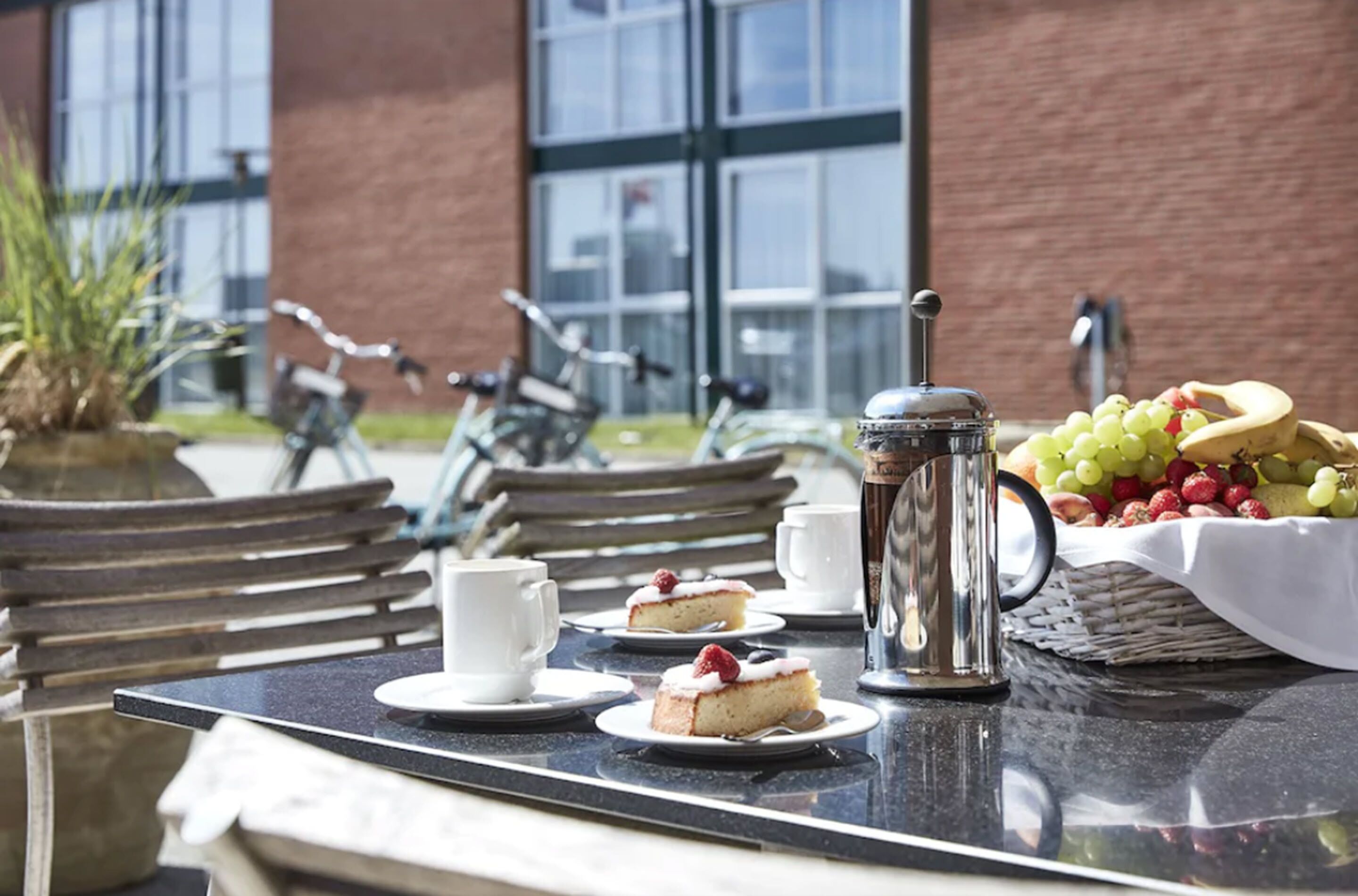 An outdoor dining area with a table set for two, including a coffee pot, cups, plates with cake and fruit, and a basket of grapes and strawberries.