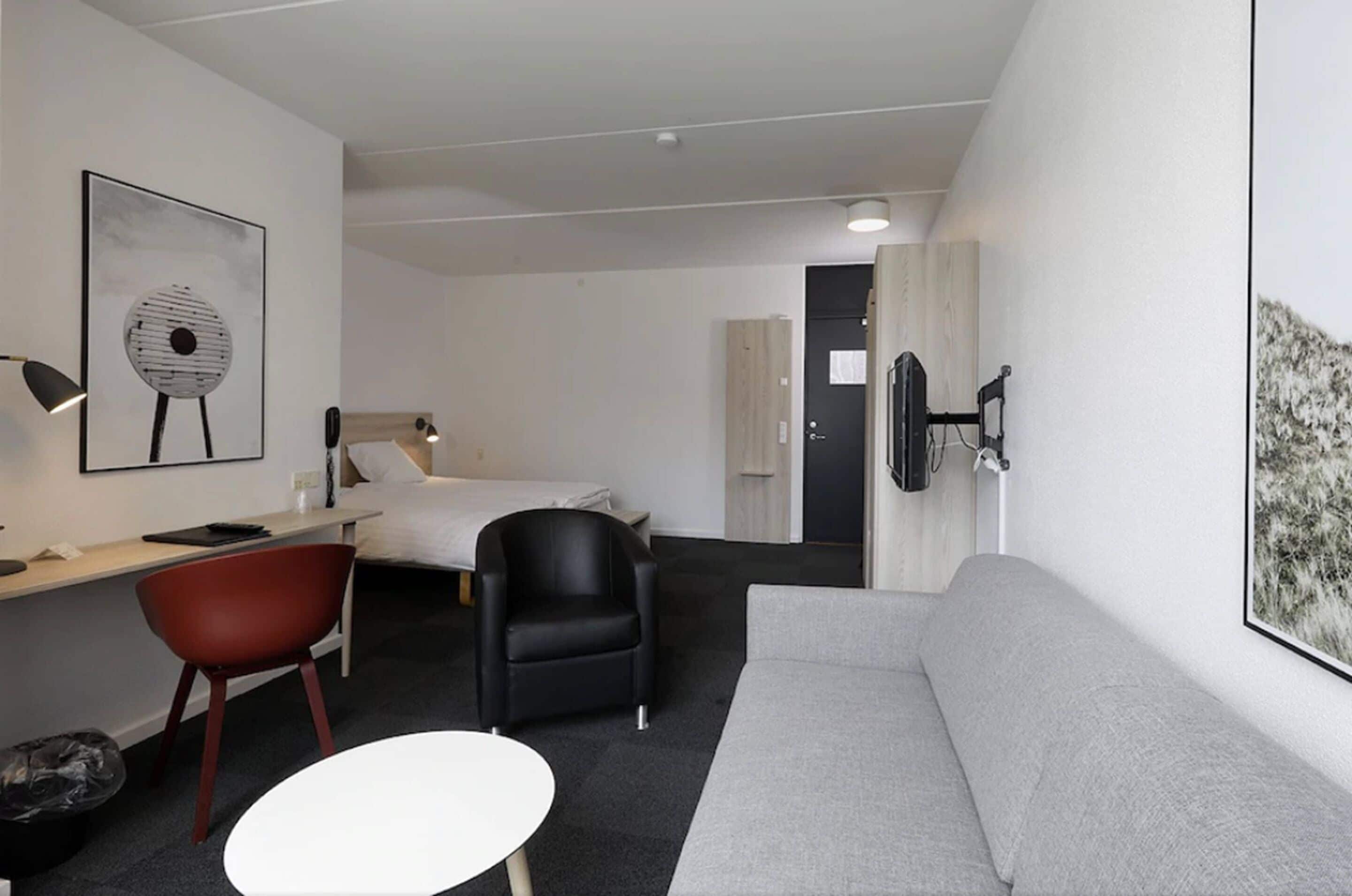 Et moderne hotelværelse med en seng, en grå sofa, en rød stol, et hvidt bord og et ophængt fjernsyn.