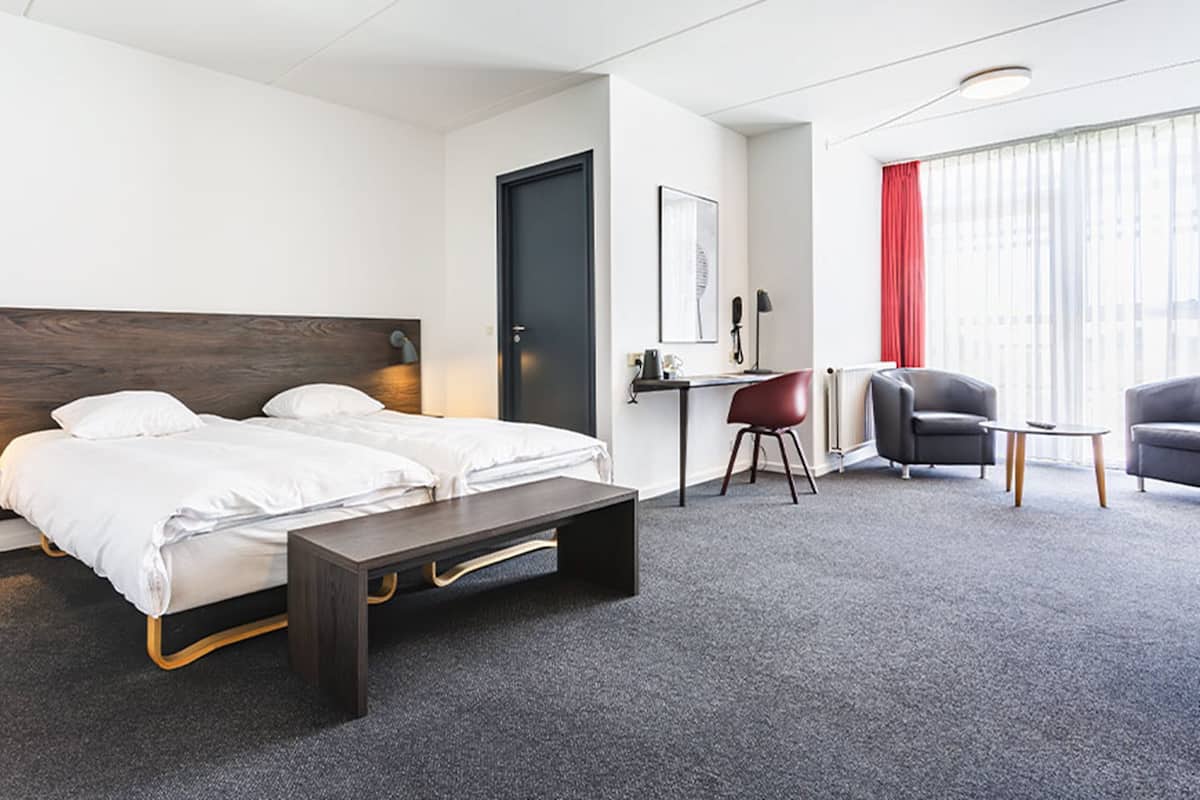 A modern hotel room with a bed, desk, two chairs, and a window with curtains.