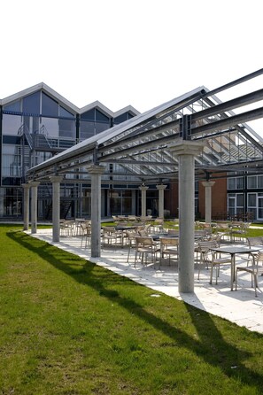 An outdoor patio area with tables and chairs, a covered walkway, and a building with large windows and a fire escape.