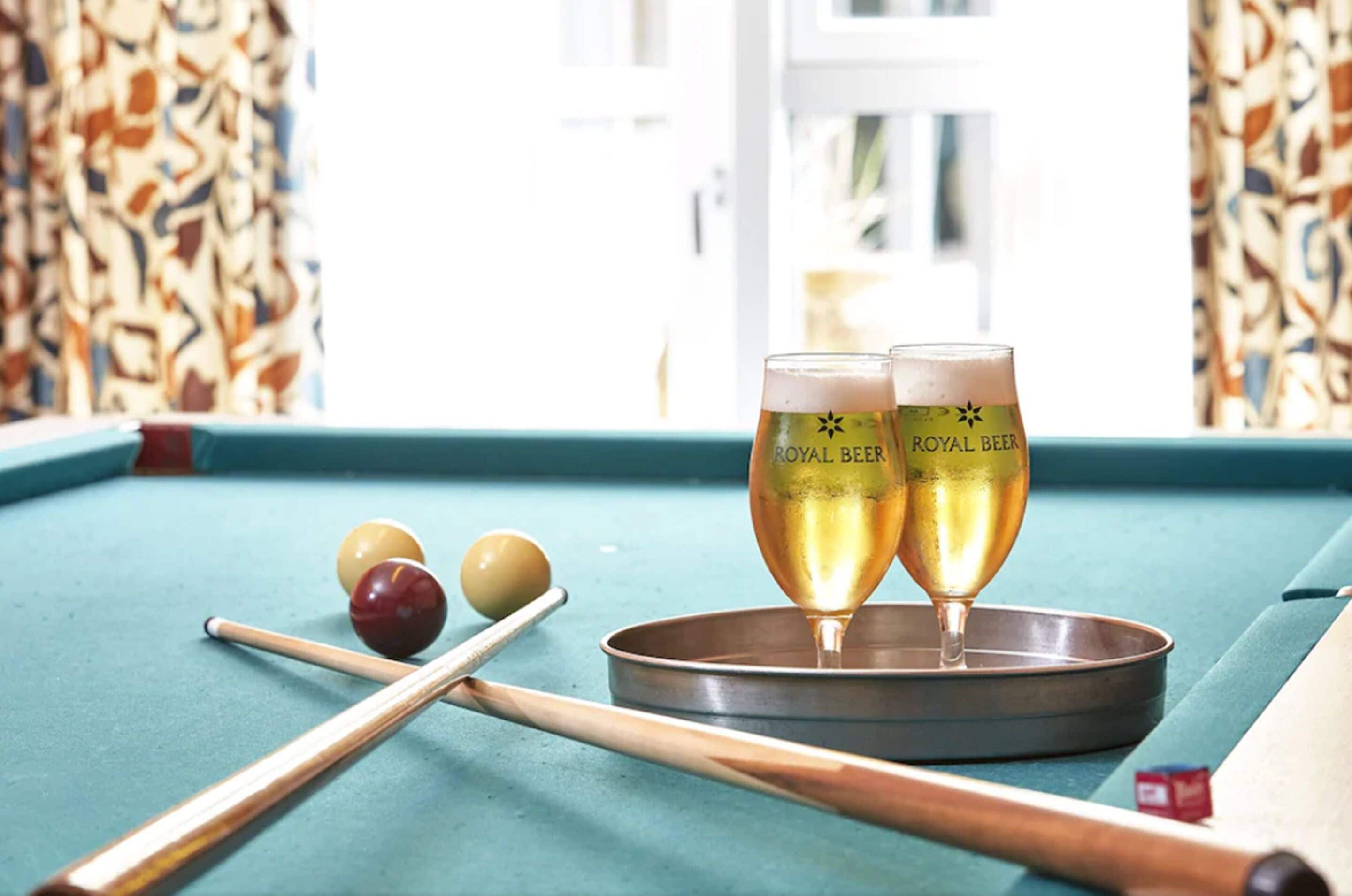 A pool table with two glasses of Royal Beer, pool cues, and billiard balls.