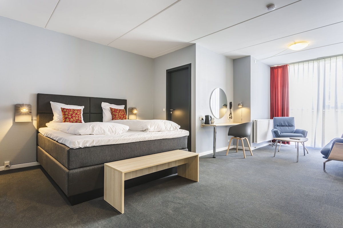 A modern hotel room with a bed, bedside table, bench, desk, chair, and a large window with curtains.