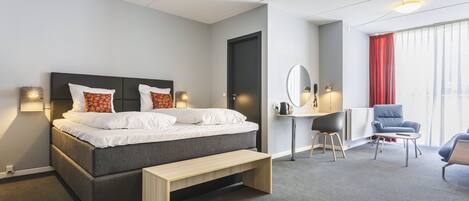 A modern hotel room with a bed, bedside table, bench, desk, chair, and a large window with curtains.