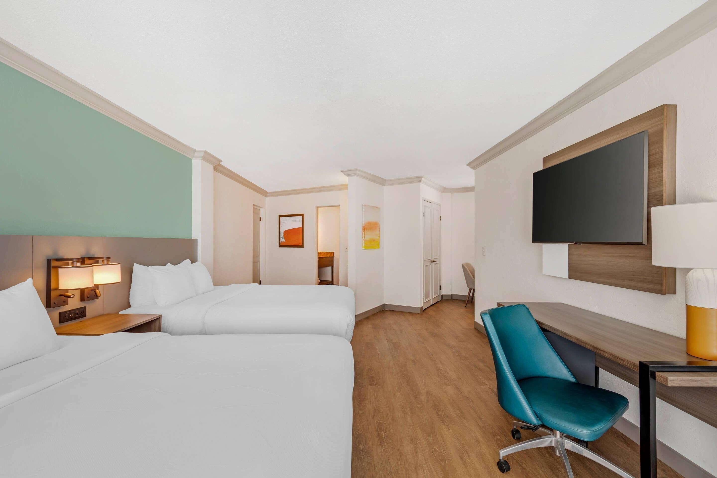 A hotel room with two beds, a desk, a chair, a TV, and a painting on the wall.