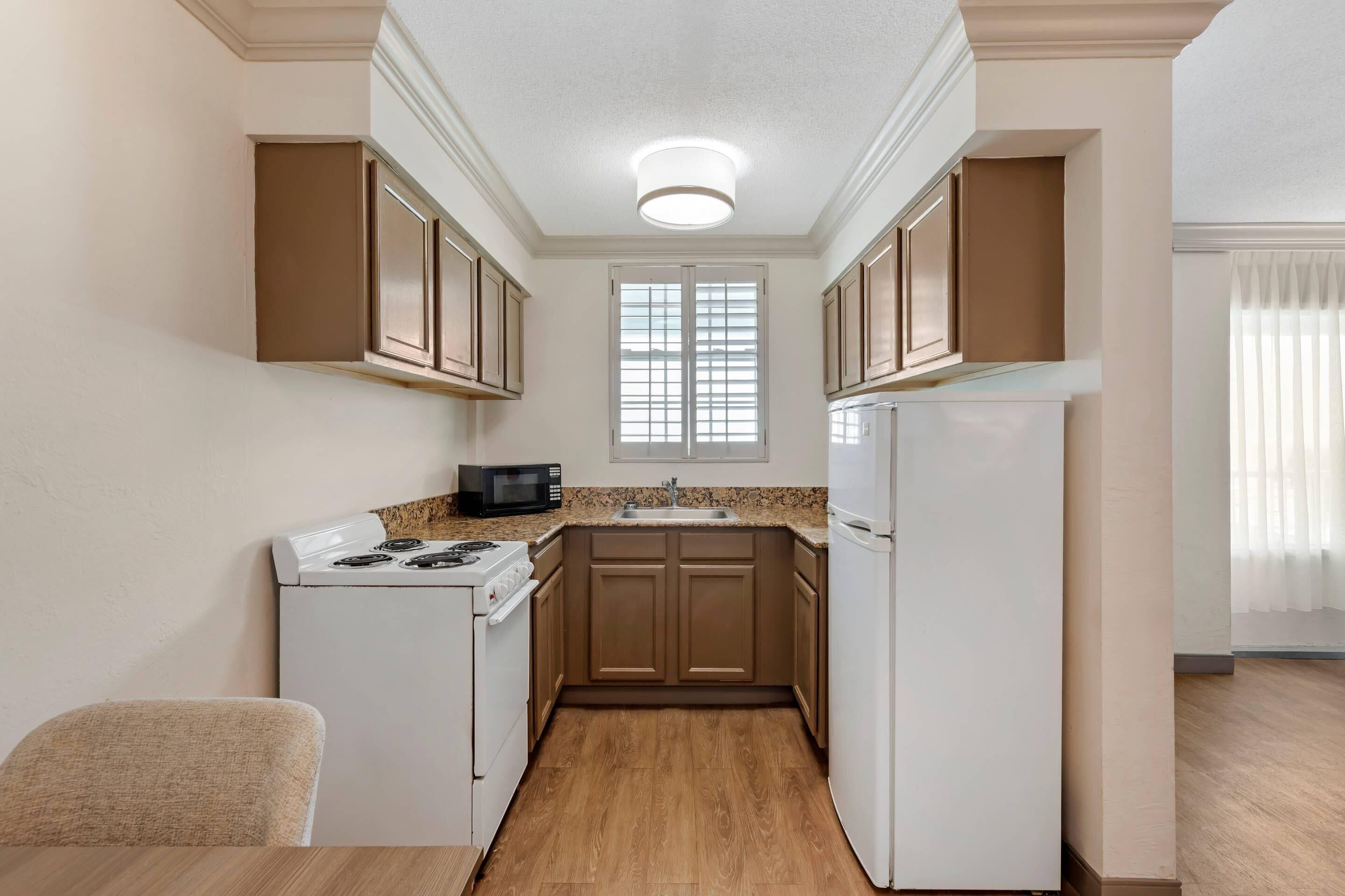 A compact kitchen with white appliances, wooden cabinets, and a window with blinds.