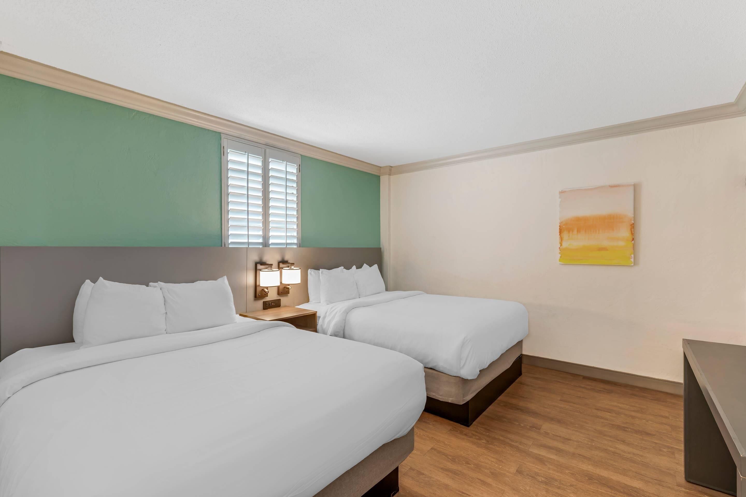 A hotel room with two beds, a window with blinds, a bedside table with lamps, and a painting on the wall.