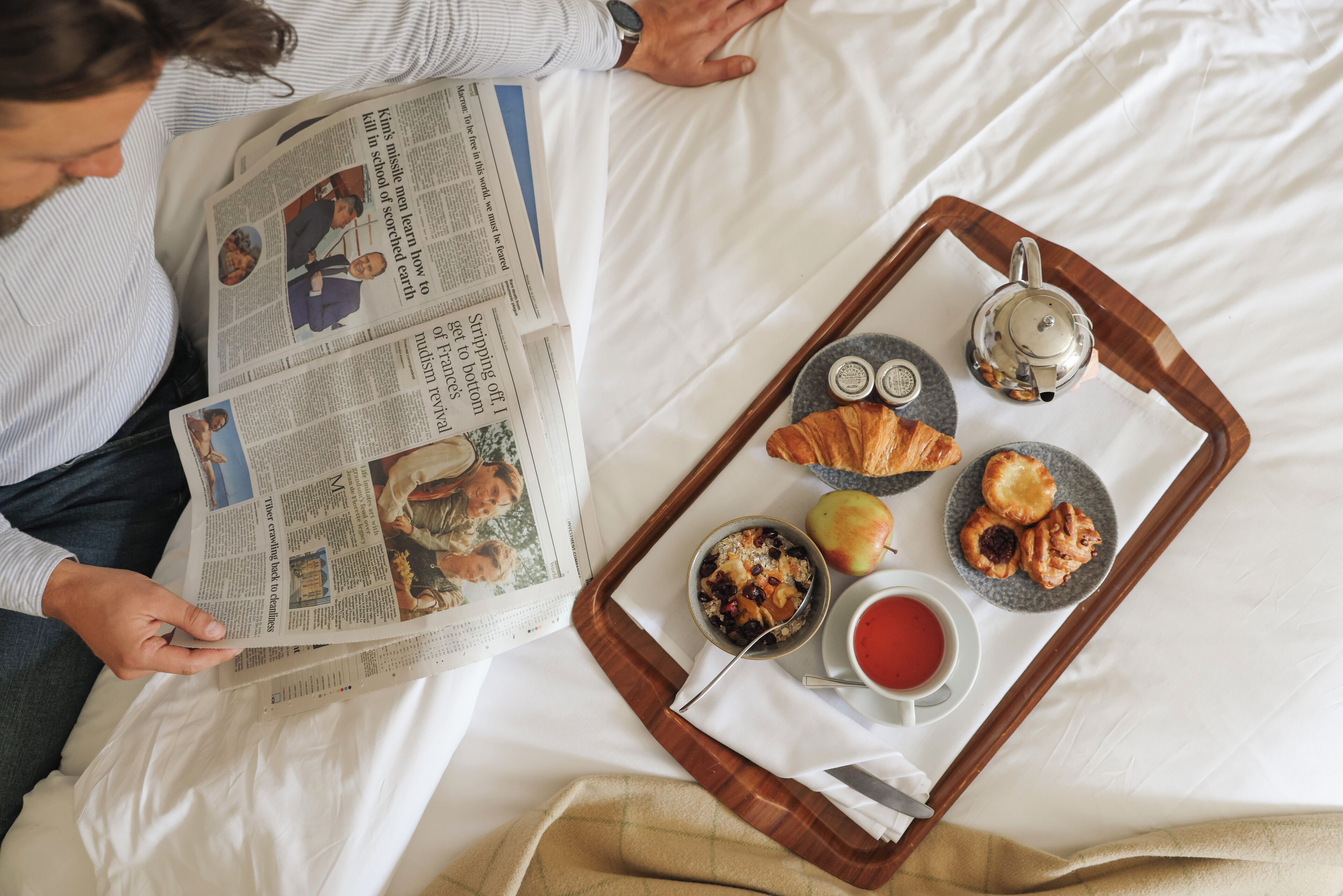 A person reading a newspaper on a bed with a tray of breakfast items including croissants, an apple, and a teapot.