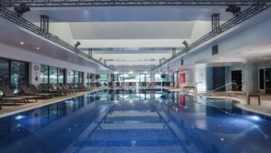 A large indoor swimming pool with a clear view of the outside, sun loungers along the sides, and a modern architectural design with a high ceiling and metal truss structure. - Donnington Hotel, Spa & Golf (Newbury)
