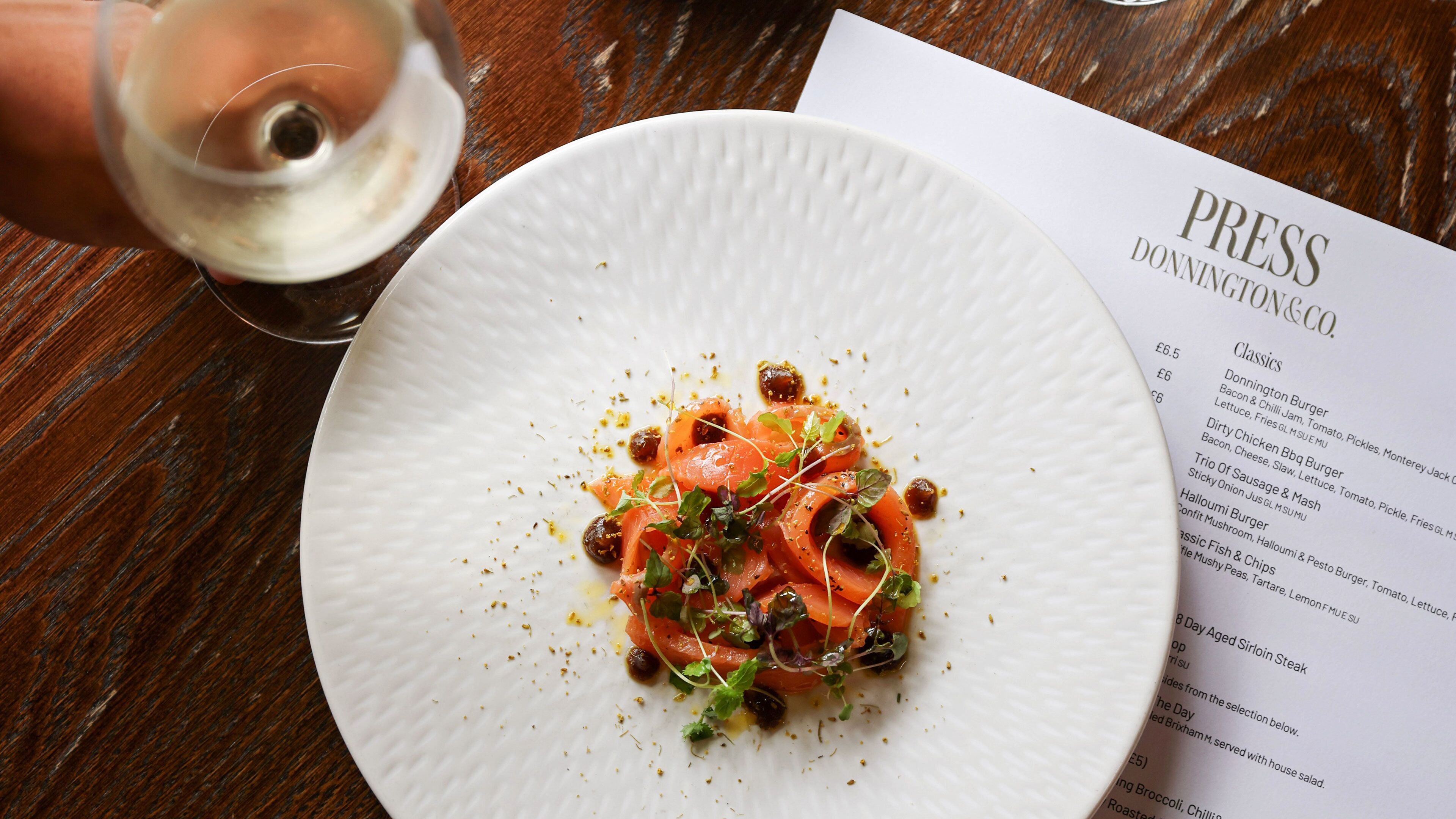 A white plate with a dish of food, a menu, and a glass of white wine on a wooden table.
