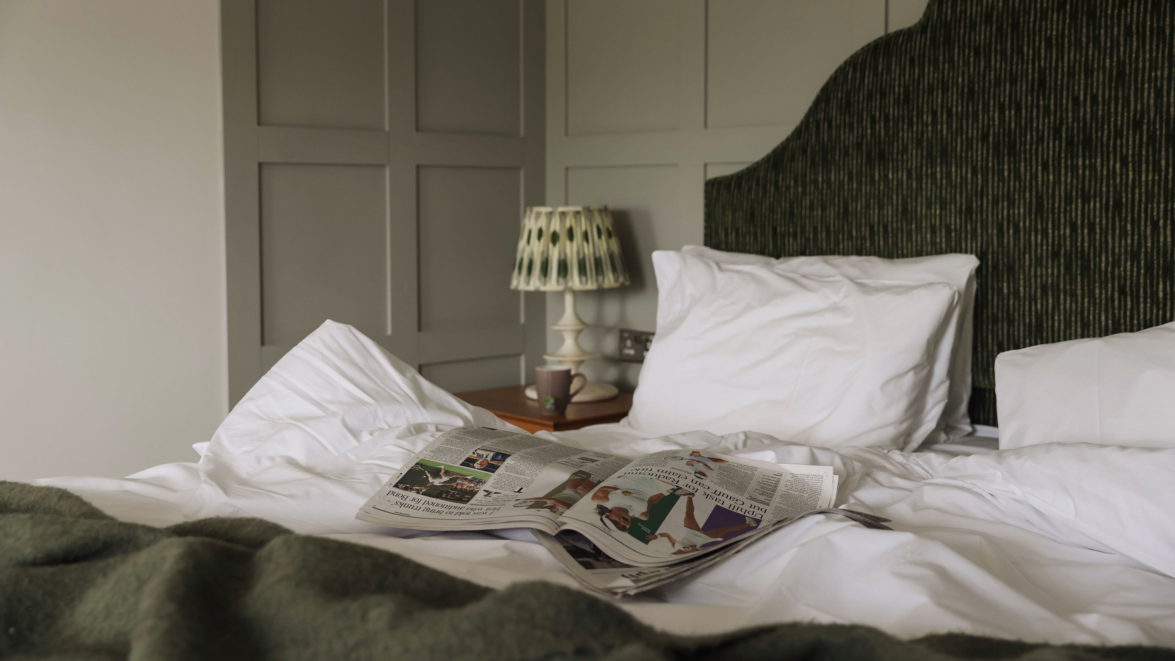 A neatly made bed with white linens, a lamp on a bedside table, and a magazine on the bed.