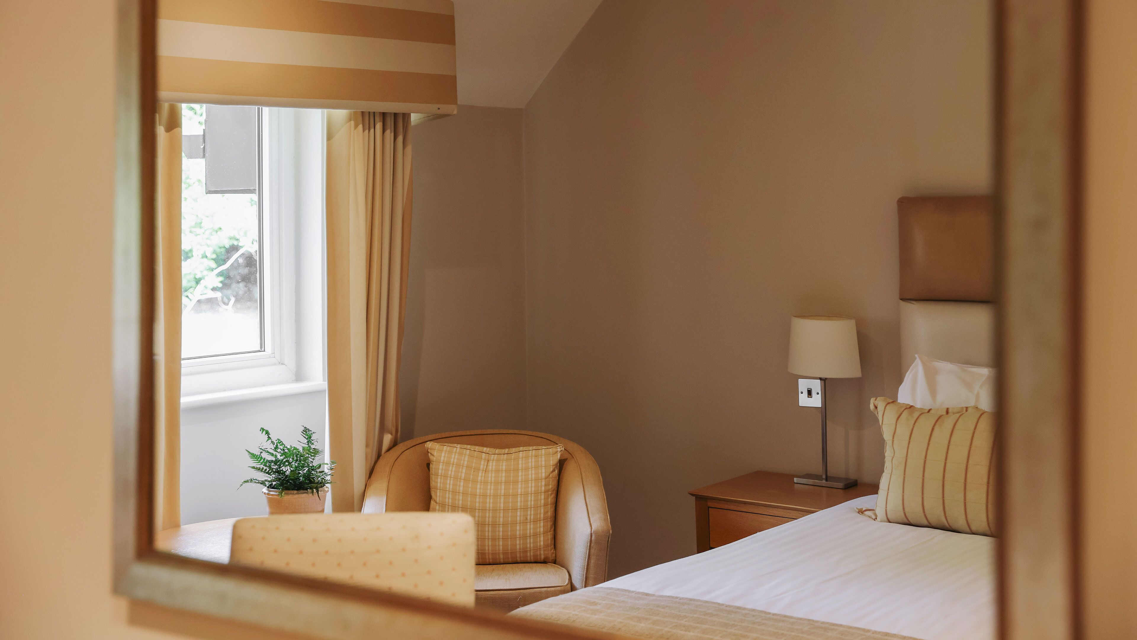 Una habitación de hotel con cama, una silla, una mesita de noche, una lámpara y una ventana con cortinas.